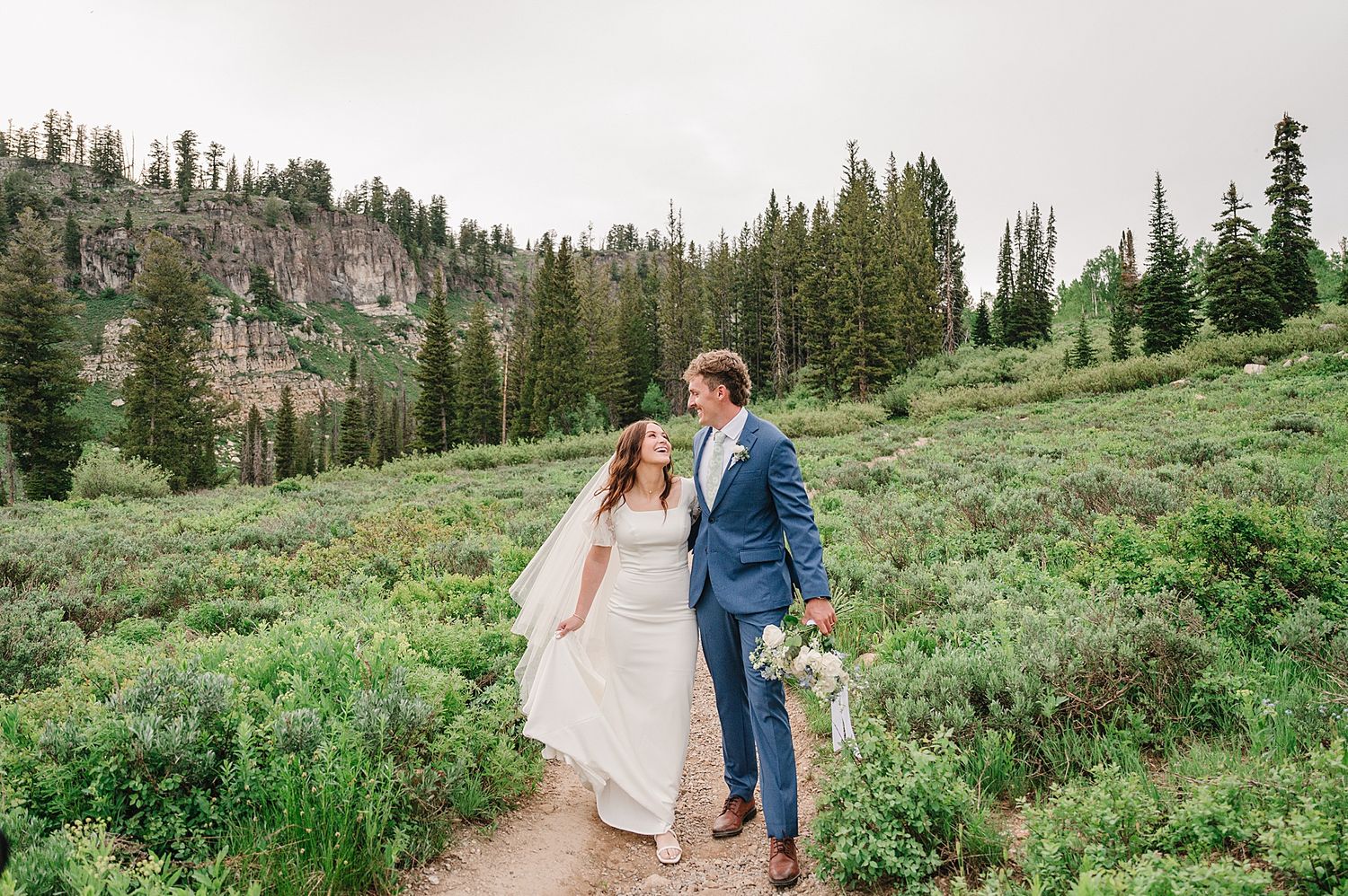 A couple walking together on a mountain trail surrounded by evergreen trees and wildflowers in a scenic mountain landscape.