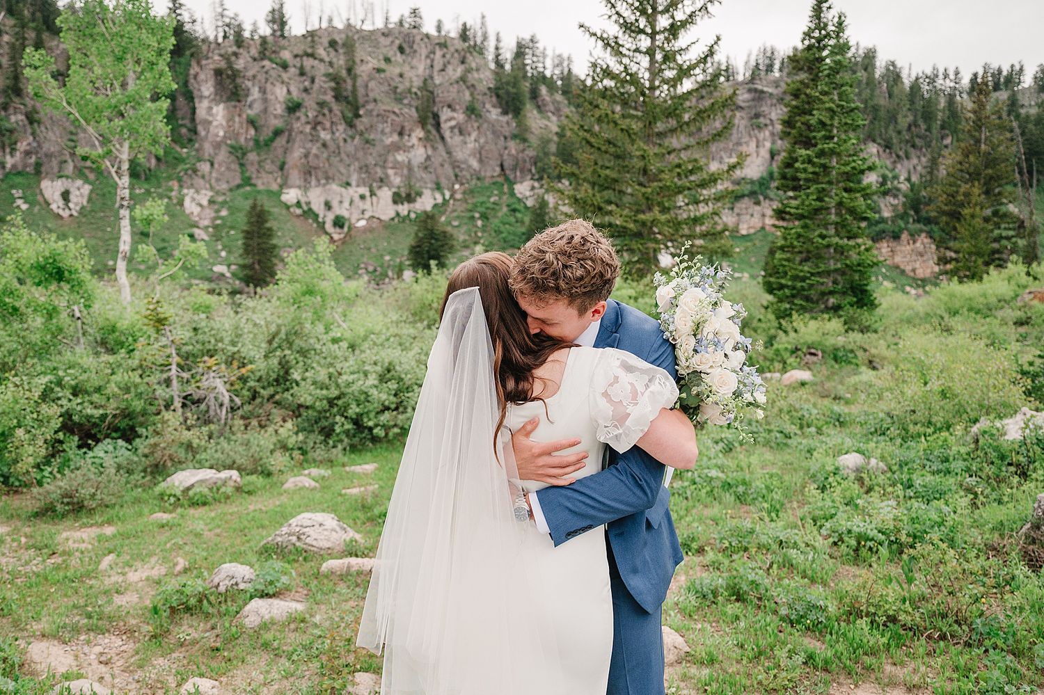 Two people share a romantic embrace in a mountain meadow surrounded by evergreen trees and rocky cliffs.