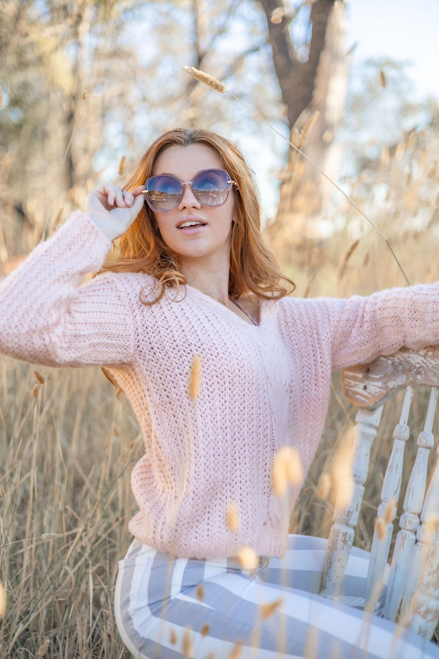 Senior portrait of girl in pink sweater and pants with blue/white stripes in a golden wheat field, posing with sunglasses