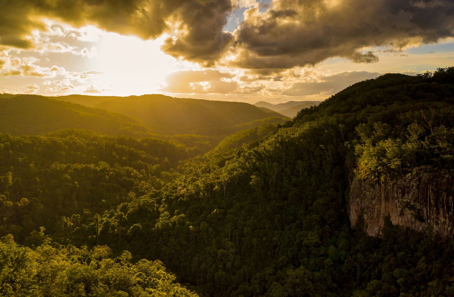 Golden sunlight breaks through storm clouds over a lush mountain valley with dramatic cliff faces.