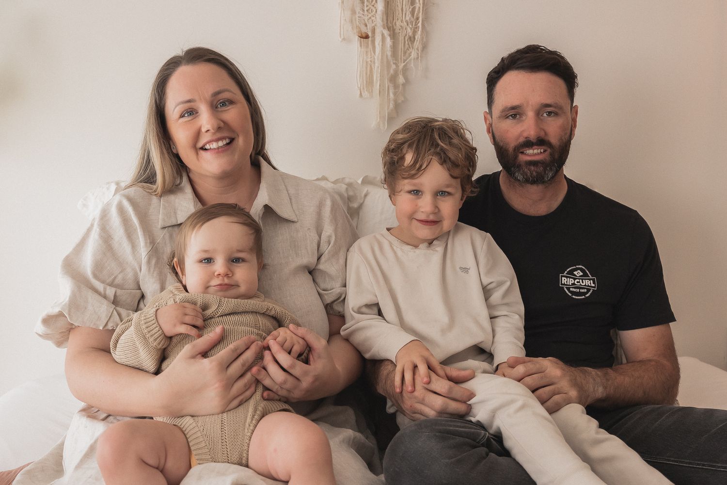 A family of four wearing beige and black clothes sitting together and smiling against a neutral background.