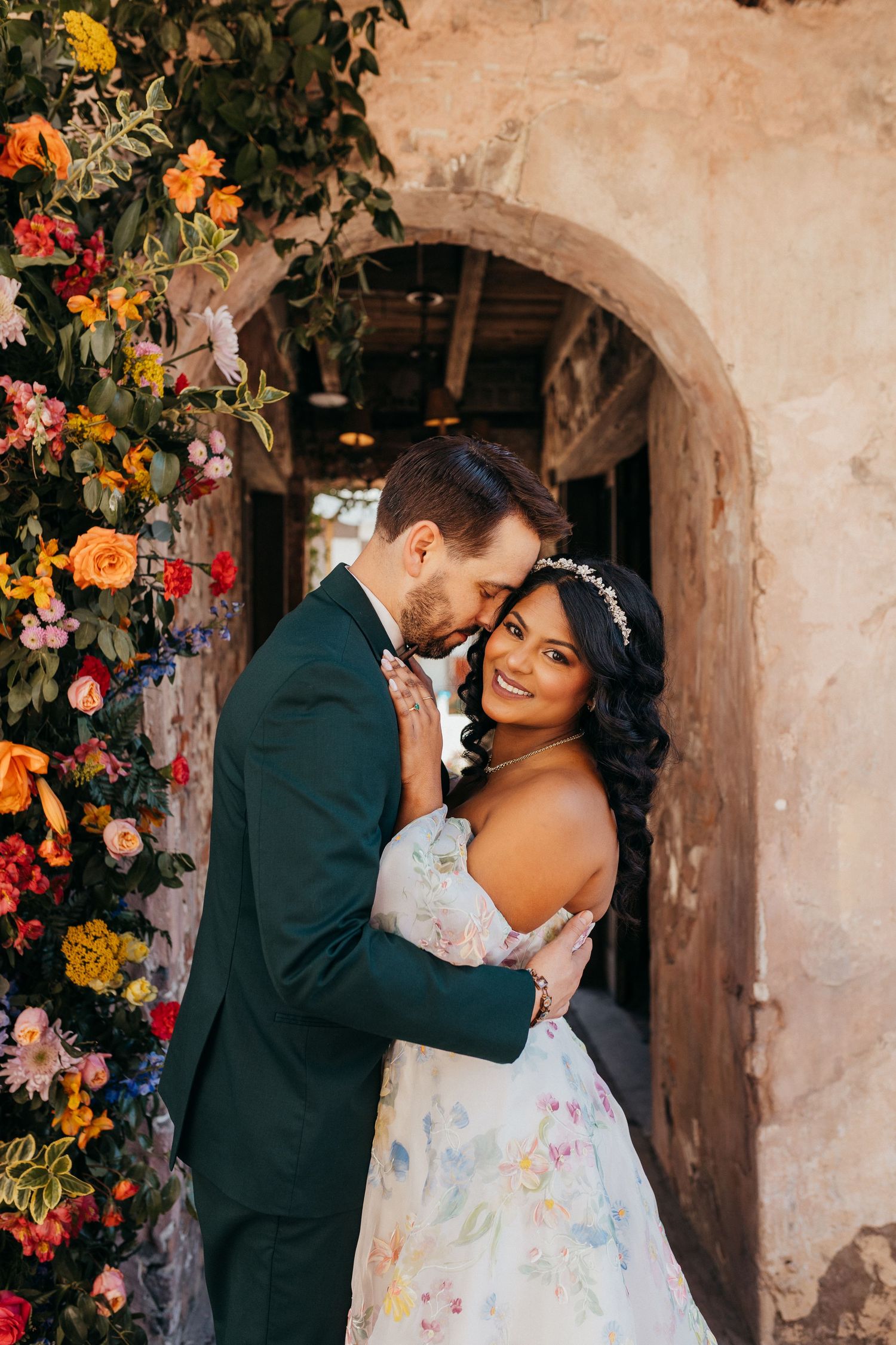 A bride in a floral print dress and groom in dark suit embrace in front of an arched stone passageway adorned with colorful flowers.