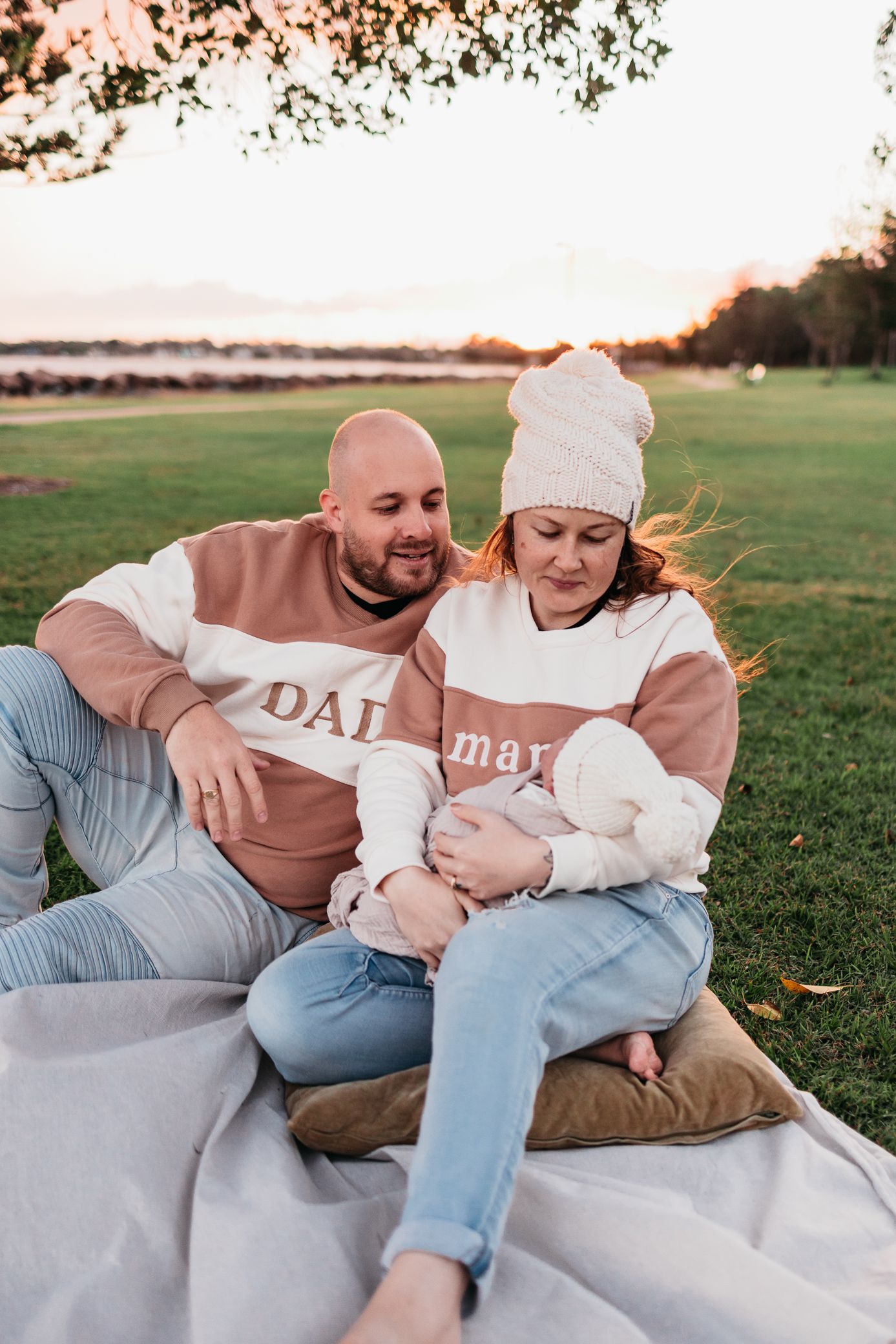 New Mum and Dad sitting on grass by water with their fresh new baby for a newborn photoshoot in Redcliffe