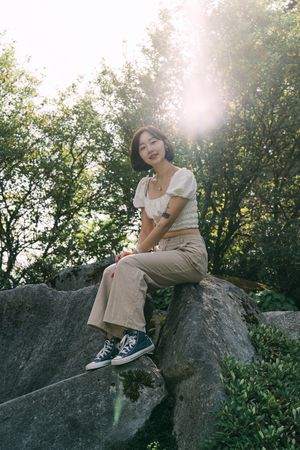 Person sitting on large boulder in sunlit forest wearing casual attire and sneakers.