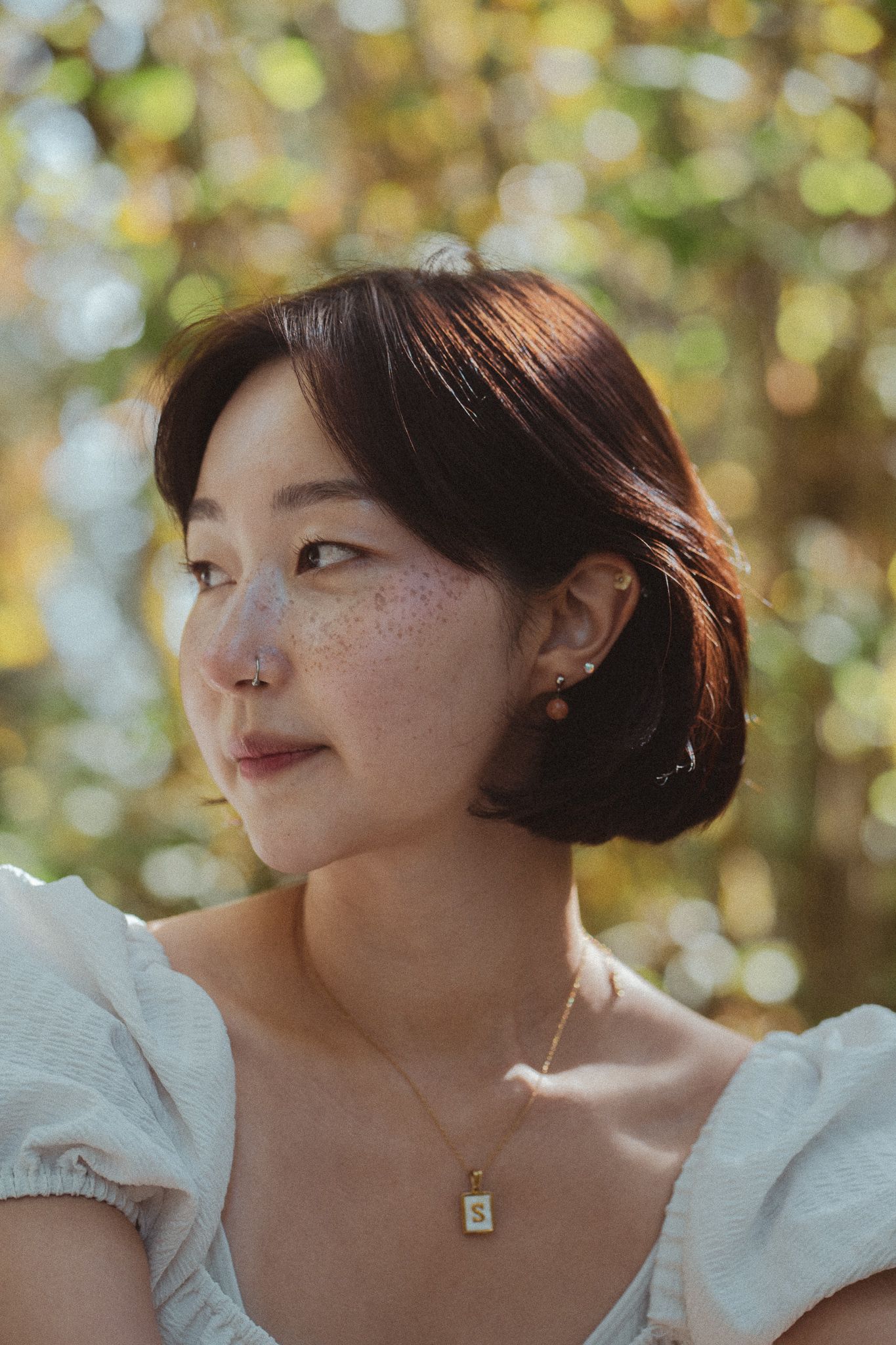 Portrait of someone with short dark hair wearing white top against blurred autumn foliage background.