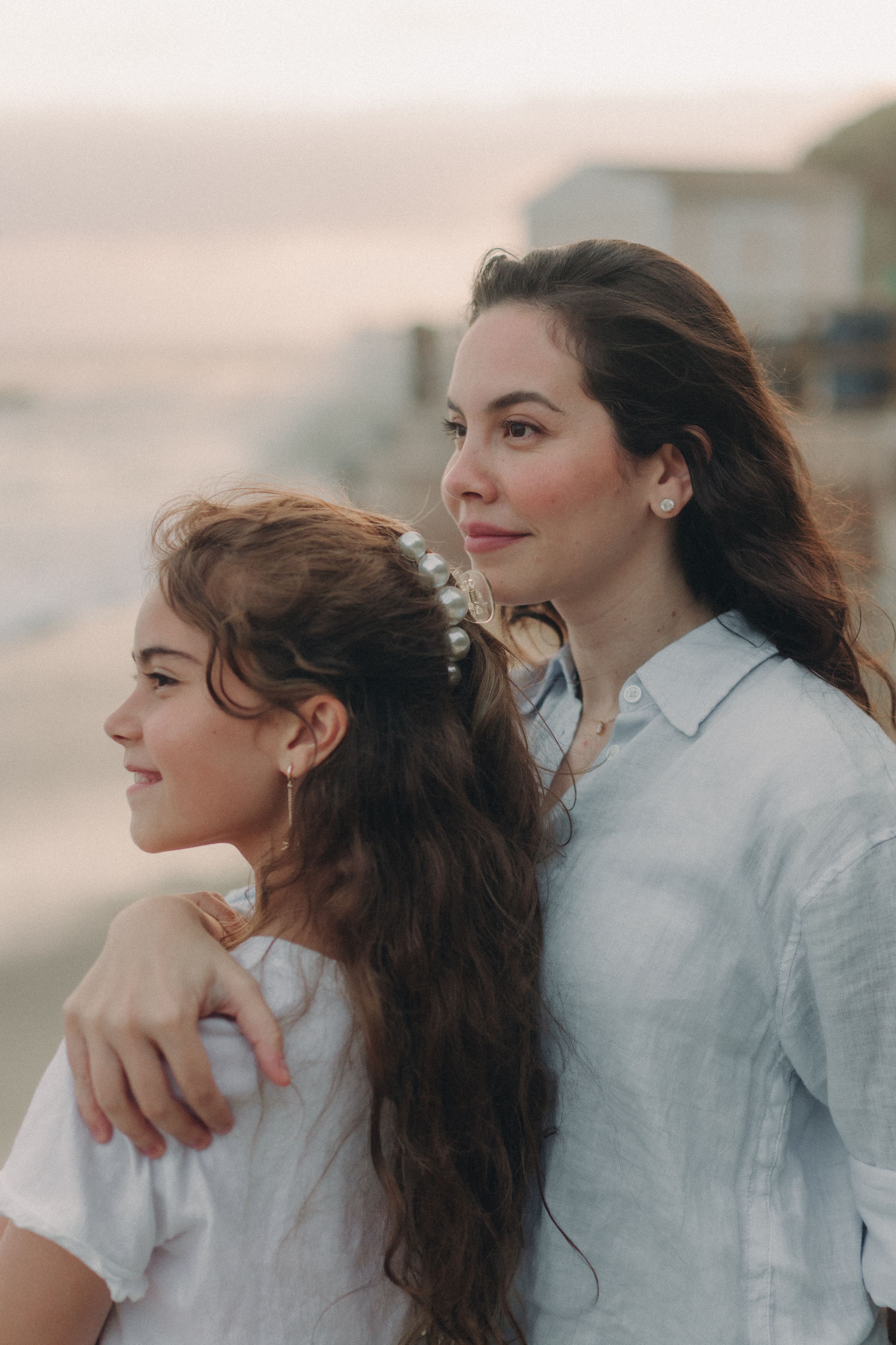 Two women with long dark hair in white shirts share an intimate moment at sunset with soft lighting.