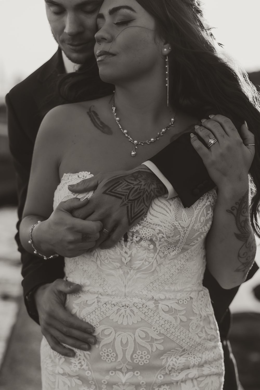 Black and white intimate wedding photo showing a romantic embrace and detailed lace dress with jewelry accents.