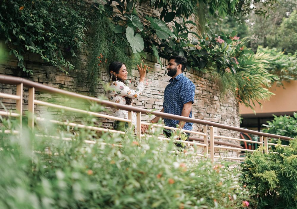Two people stand on a wooden walkway surrounded by lush green foliage in a garden setting.