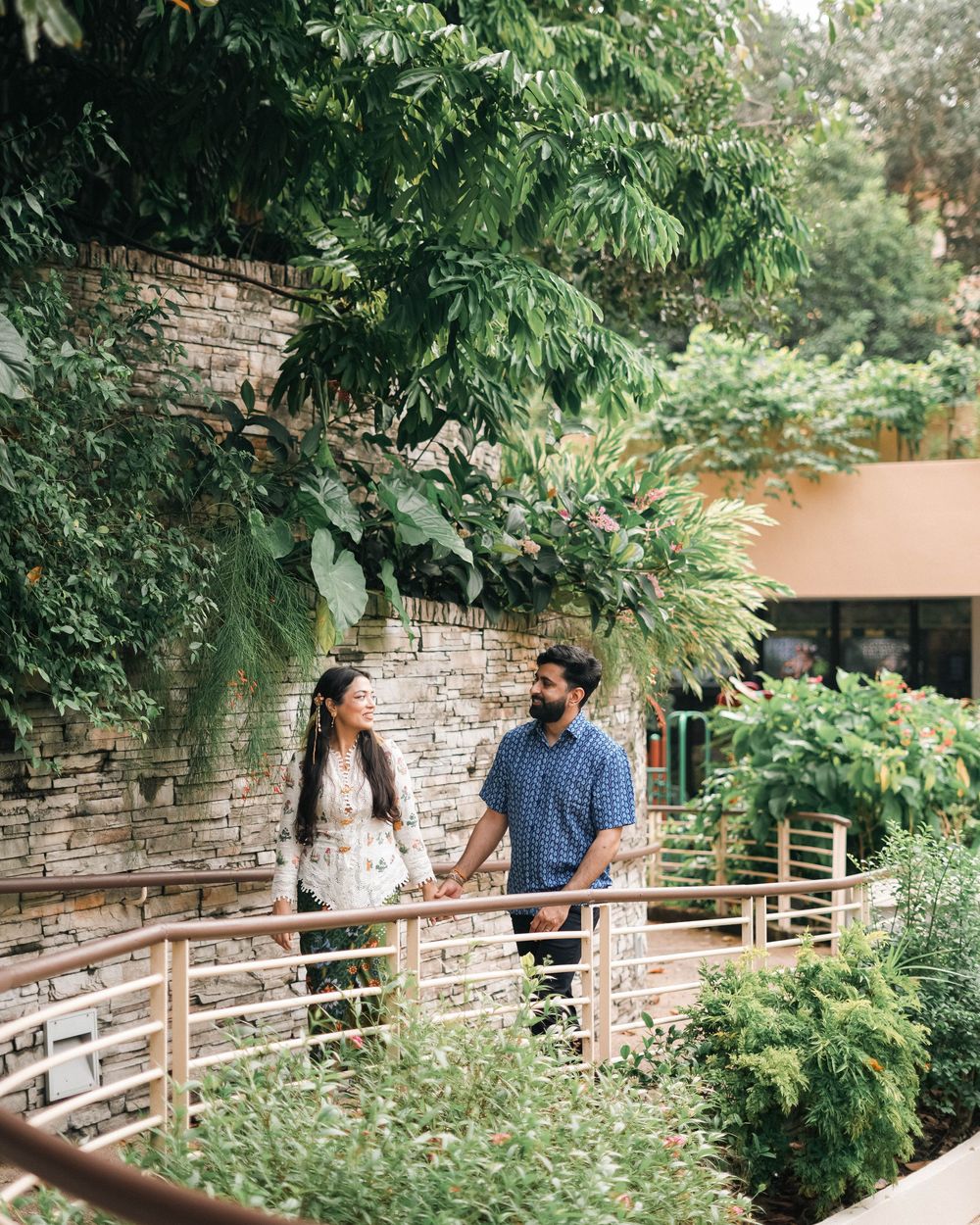 A couple stands together on a wooden walkway surrounded by lush greenery and a stone wall in a garden setting.