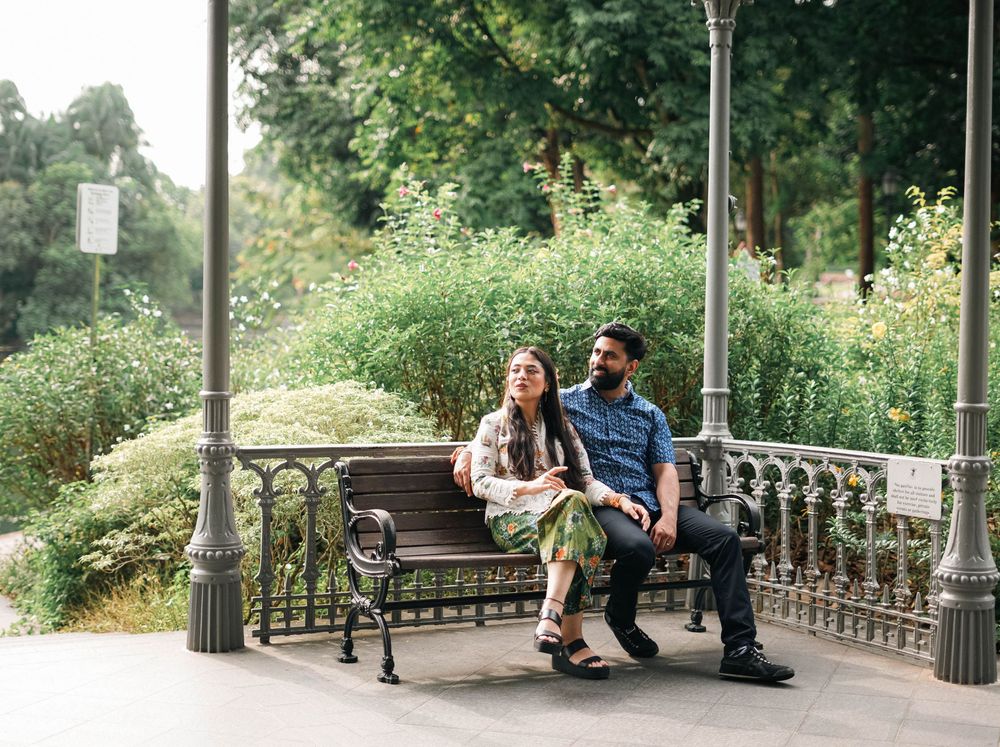 A couple sits together on an ornate bench in a park surrounded by lush greenery and decorative metal railings.