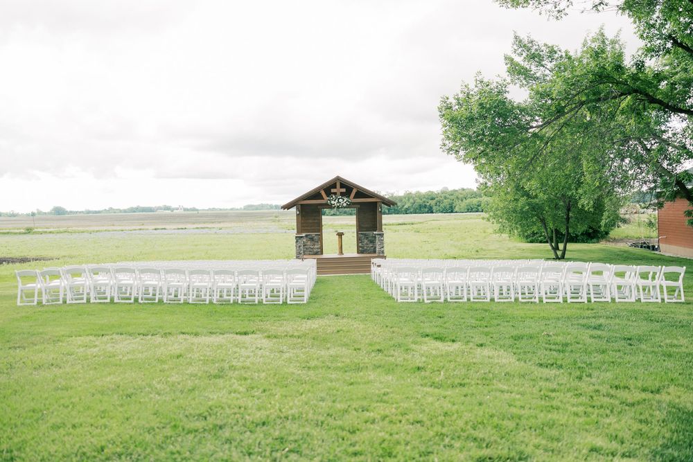 White folding chairs arranged in rows on a grassy field with a rustic building and trees in the backdrop.