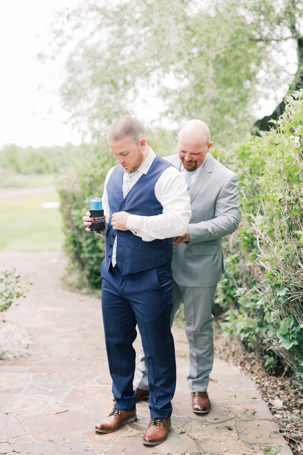 Two groomsmen in suits walking together and sharing a laugh on a garden path.