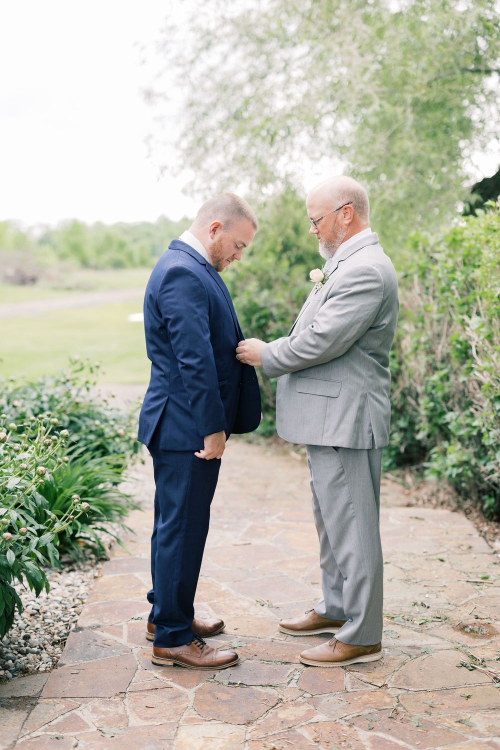 Two gentlemen in suits share an intimate moment on a garden path during what appears to be a wedding ceremony.