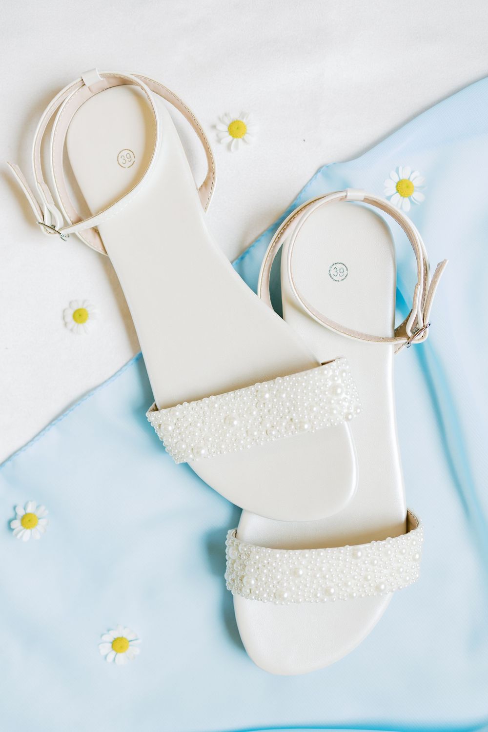 Elegant white wedding sandals with pearl details on a light blue background with tiny white flowers.