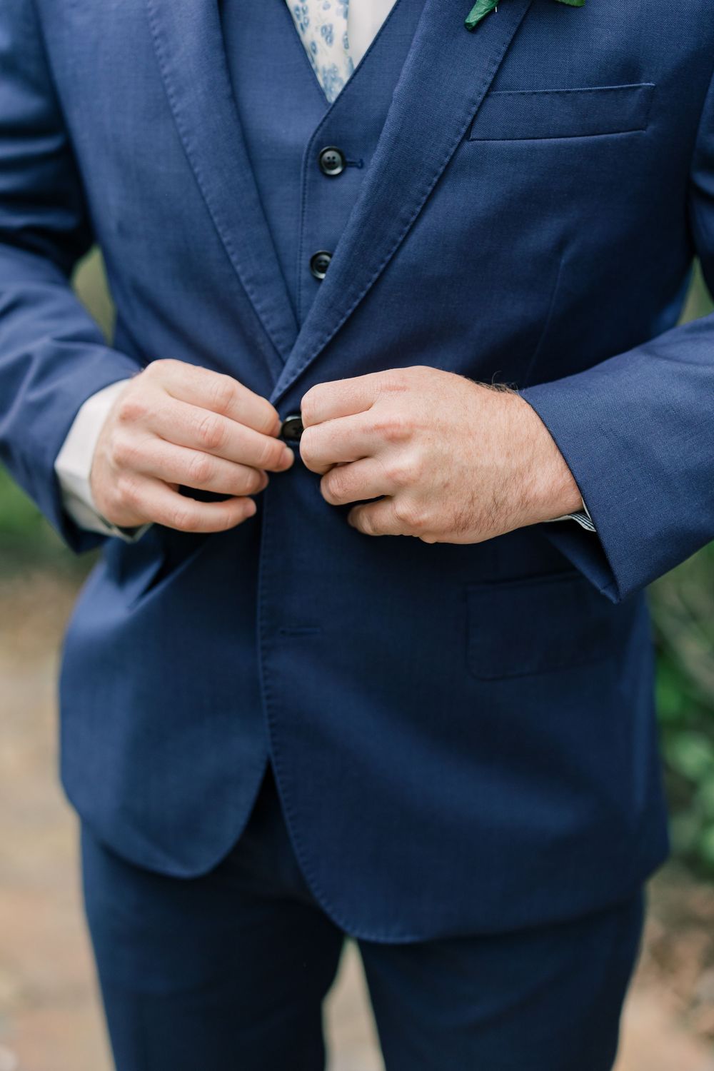 Close-up of hands buttoning a navy blue three-piece suit jacket.