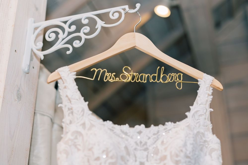 White wedding dress hanging on a personalized gold wire hanger that reads 'Mrs. Snandboy' mounted on an ornate wall bracket.