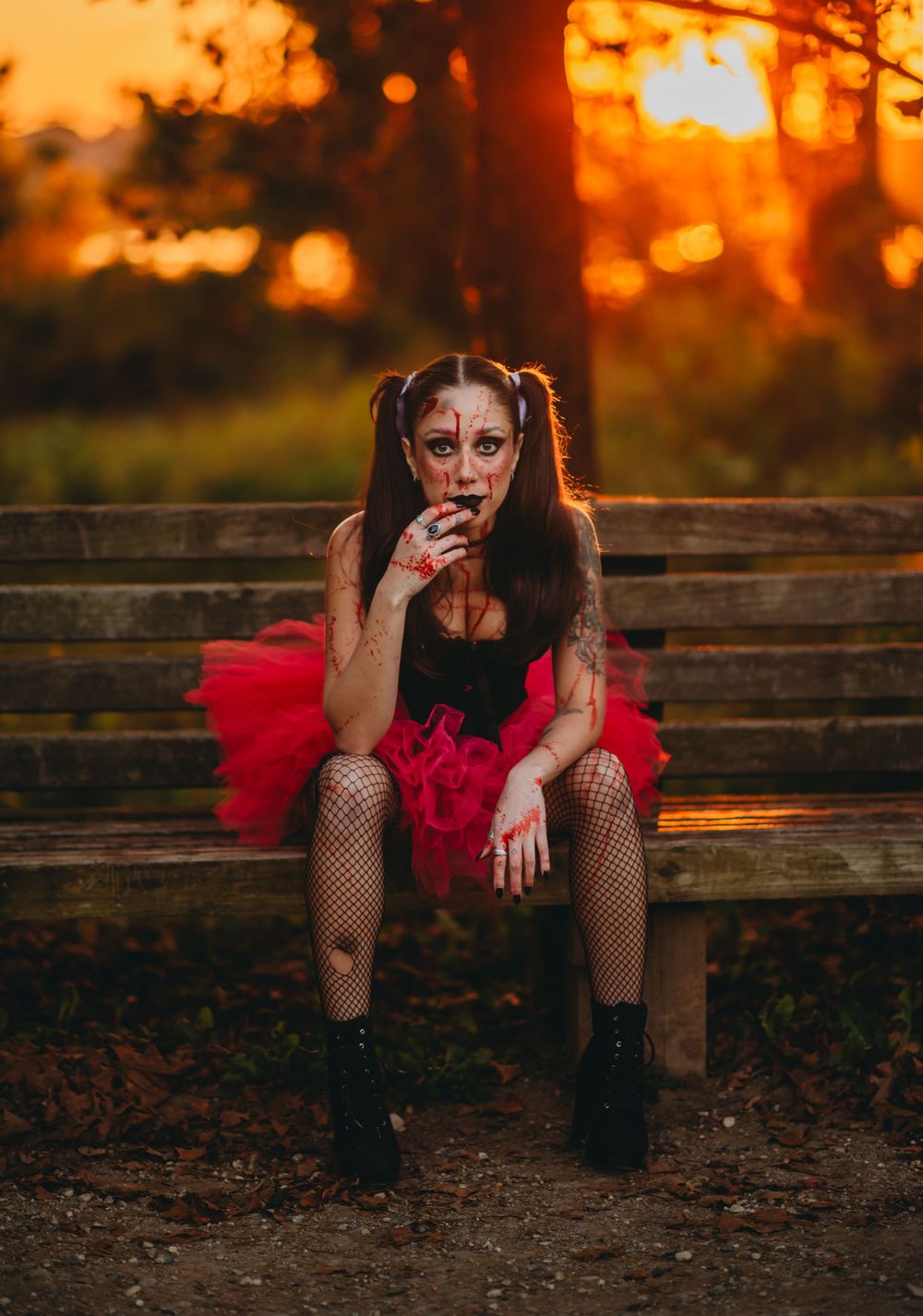 Figure in a red tutu and fishnet stockings sits on wooden bench at sunset with dramatic lighting and dark makeup.