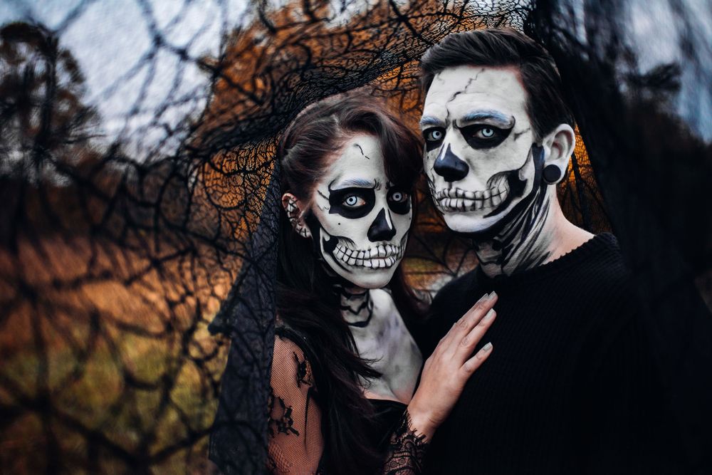 A gothic couple in dark attire wearing black and white skull face paint poses beneath a lacy backdrop during autumn.