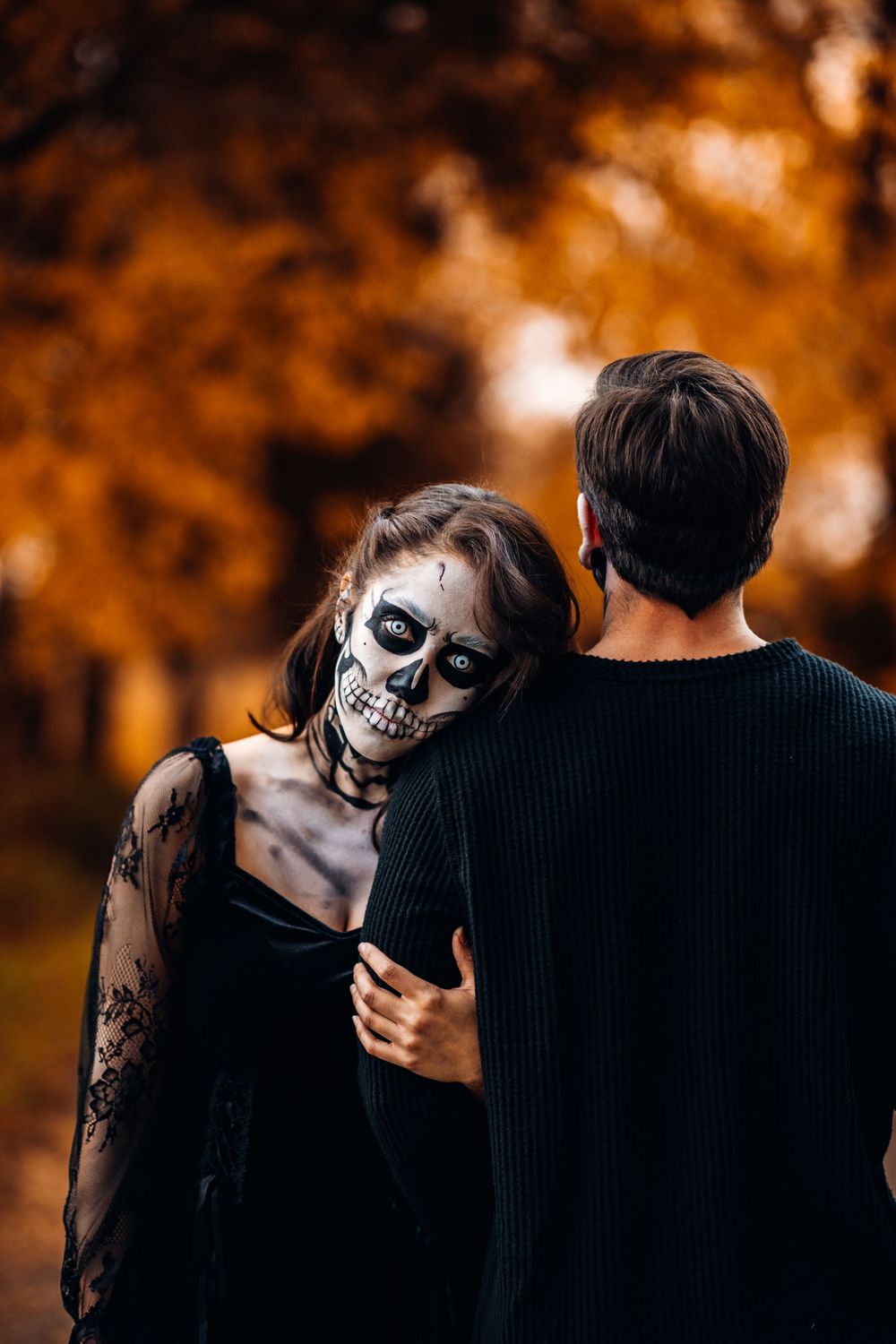 A moody couple photoshoot with Halloween-inspired skeleton face paint in an autumn forest at golden hour.
