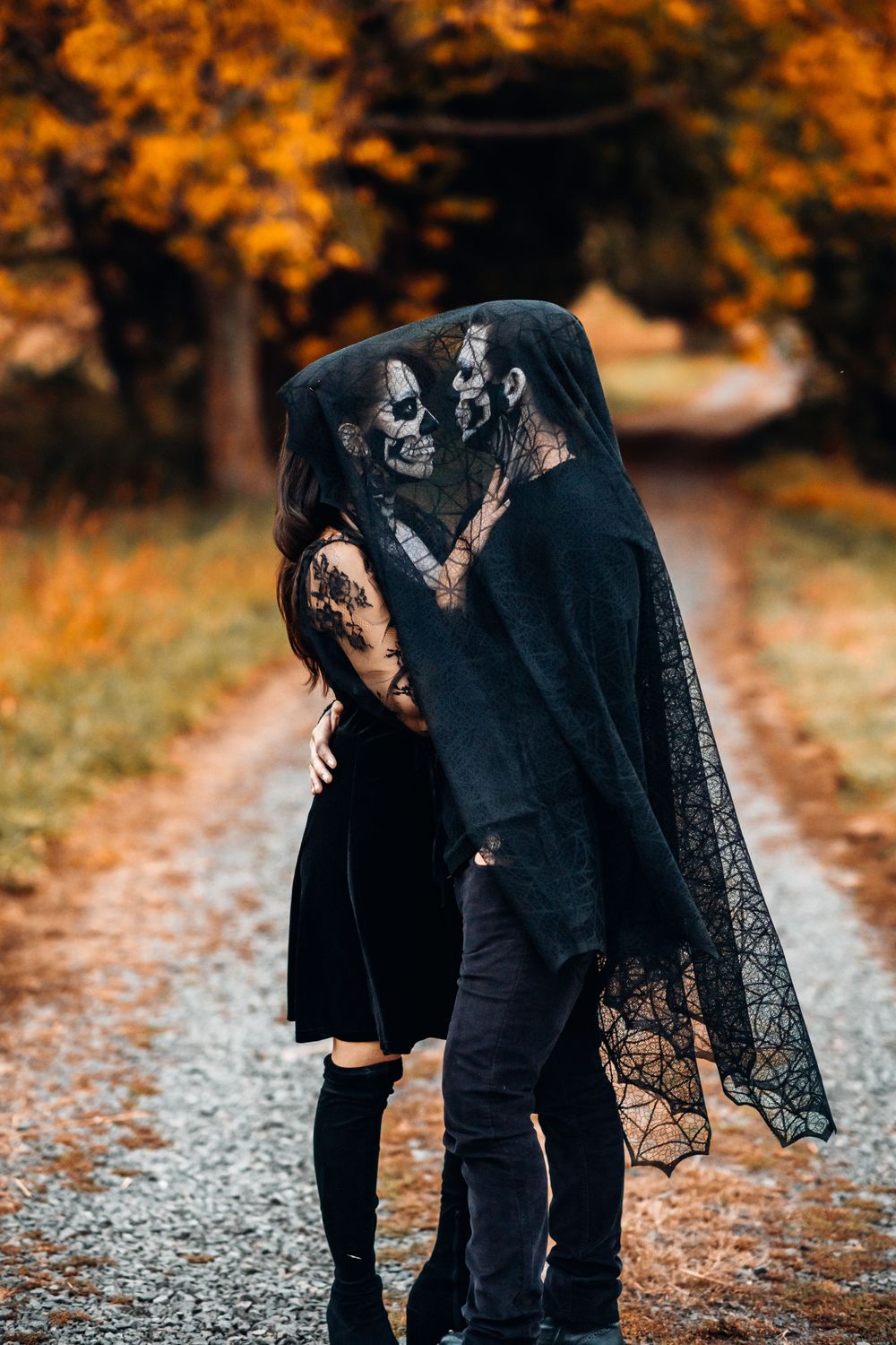 A couple in dark gothic clothing share a romantic moment on an autumn path with orange foliage in the background.