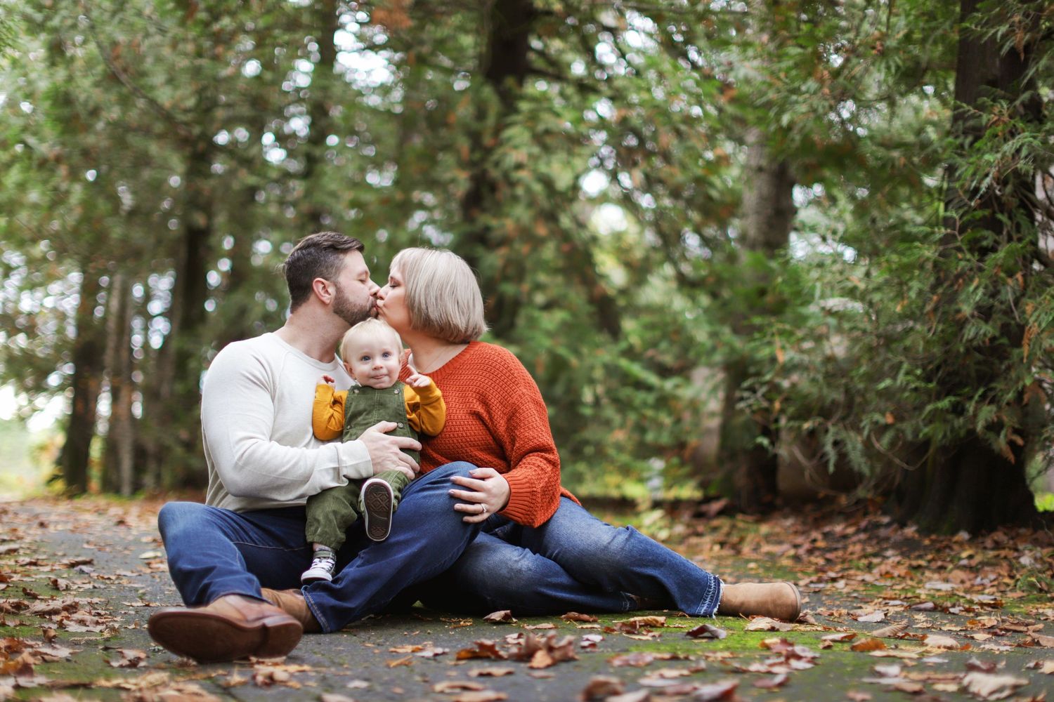 Three people in casual clothing laugh together while sitting on a forest slope surrounded by tall pine trees.