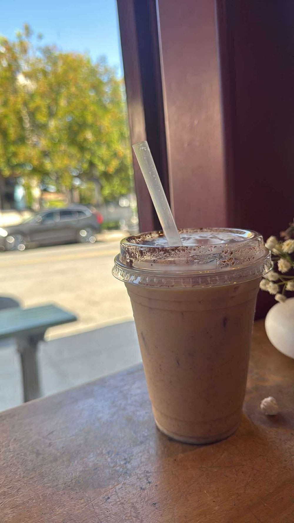 A chocolate iced coffee drink in a clear plastic cup sits on a brown table by a sunny window.