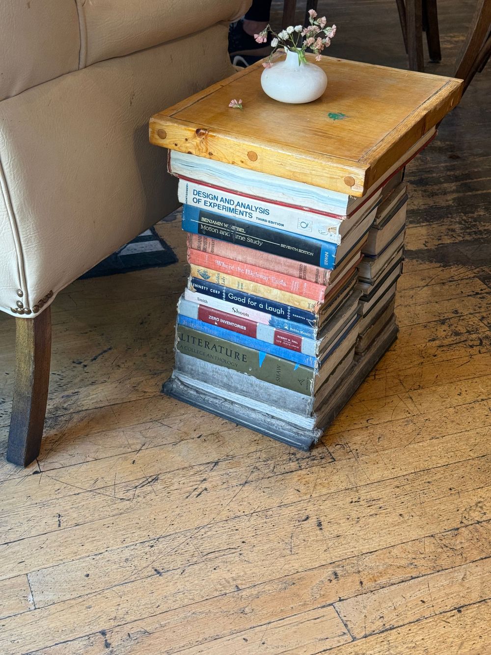 Creative side table made from stacked books topped with a wooden surface and small white vase with flowers.
