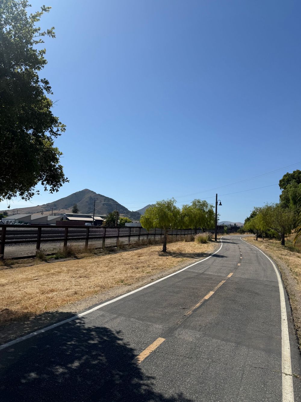 A paved bike path curves along a rural road with mountains visible in the distance under clear blue skies.