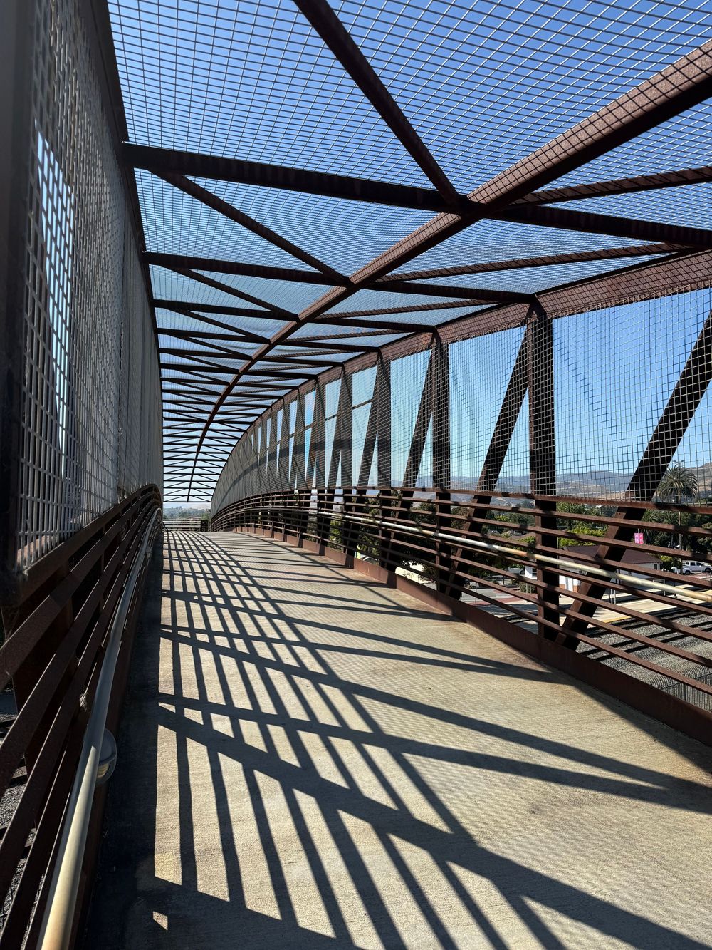 Metal pedestrian bridge with geometric support beams casting dramatic shadows through safety netting on a sunny day.