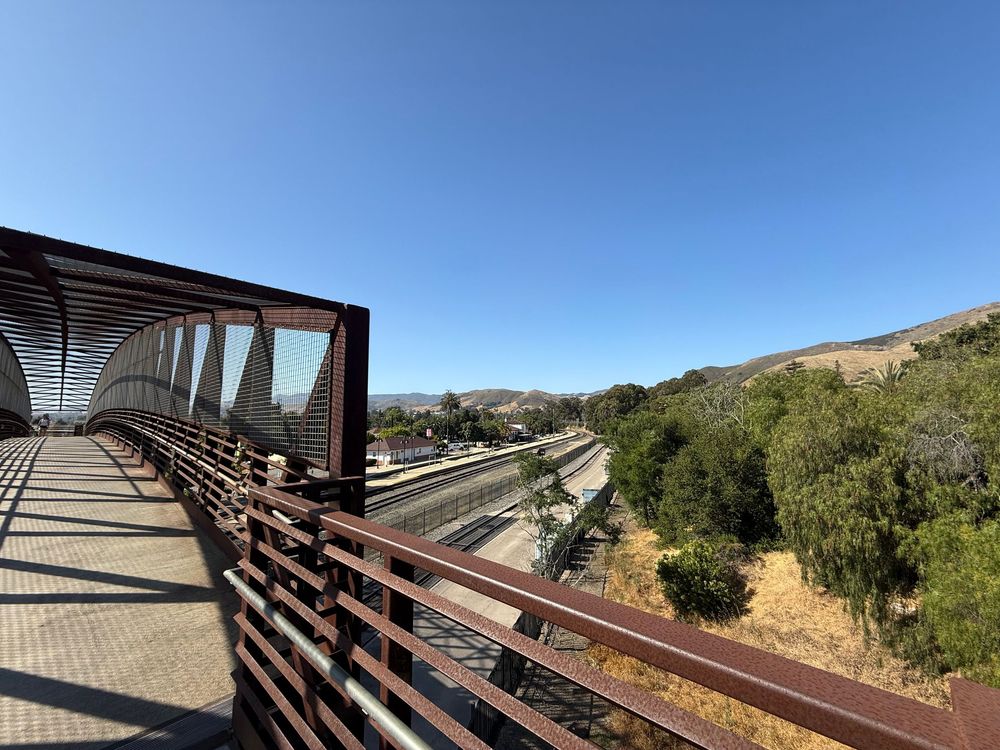 Covered pedestrian bridge spans over highway with mountains and blue sky in Southern California landscape.