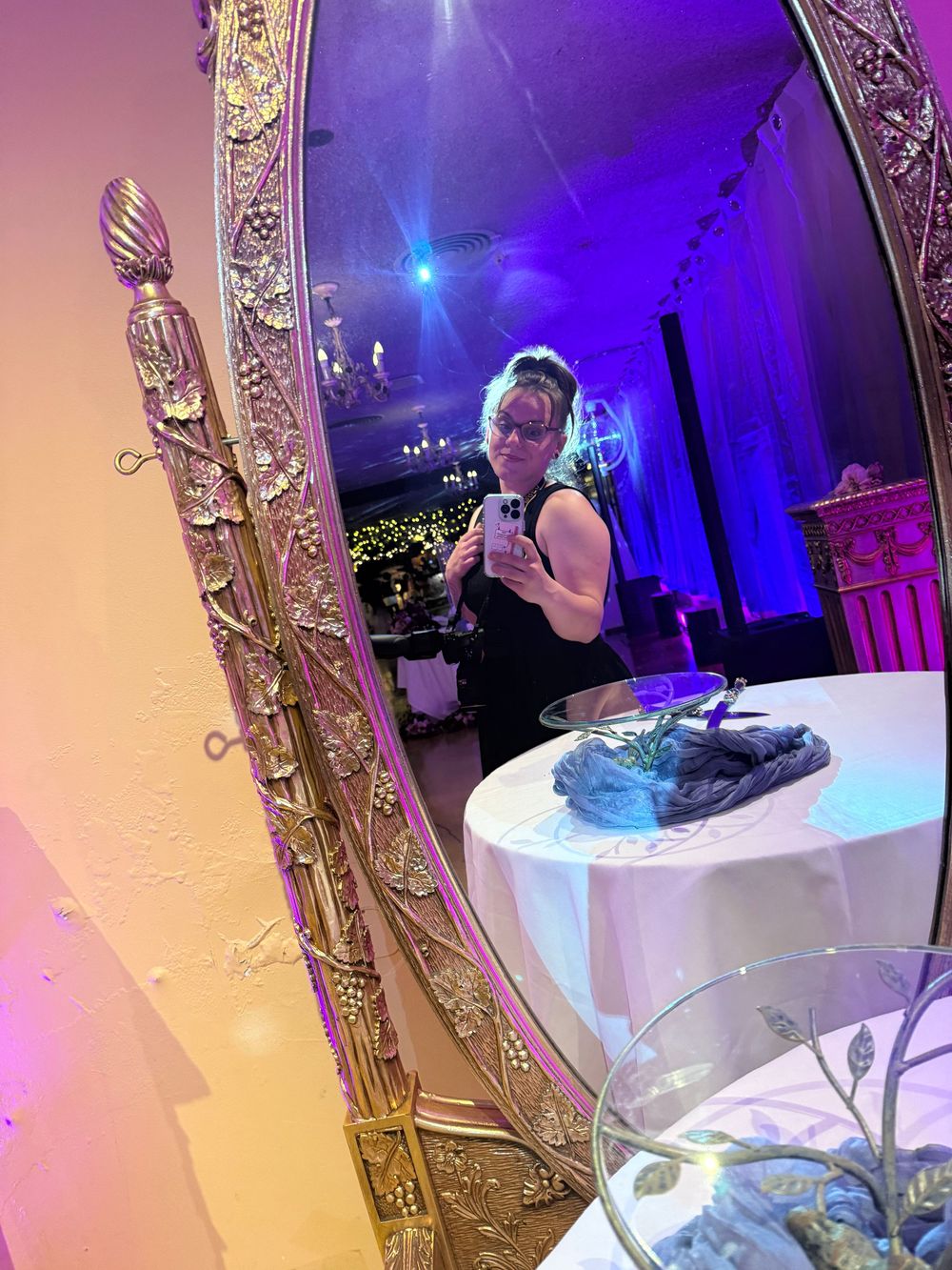 Ornate gold framed mirror reflects a dimly lit room with purple and blue ambient lighting and elegant party decor.