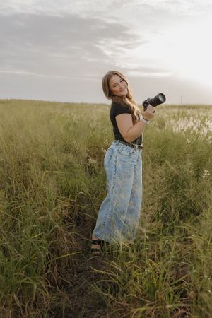A photographer in denim overalls poses playfully in a golden grass field at sunset.