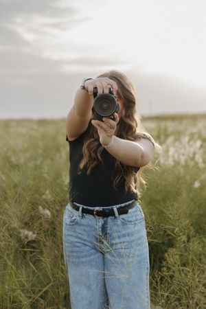 A photographer wearing jeans and black top takes photos in a tall grass meadow.