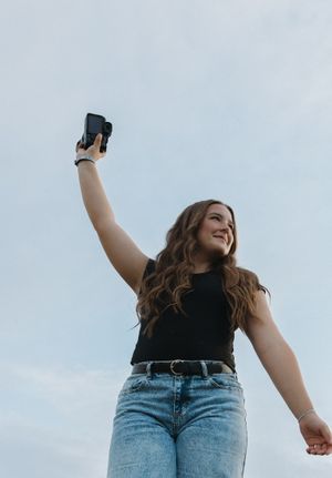 Photographer holding up a camera against cloudy sky while wearing black top and jeans.