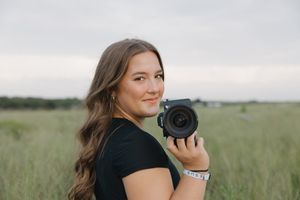 Portrait of photographer with long wavy hair holding camera in grassy field at dusk.