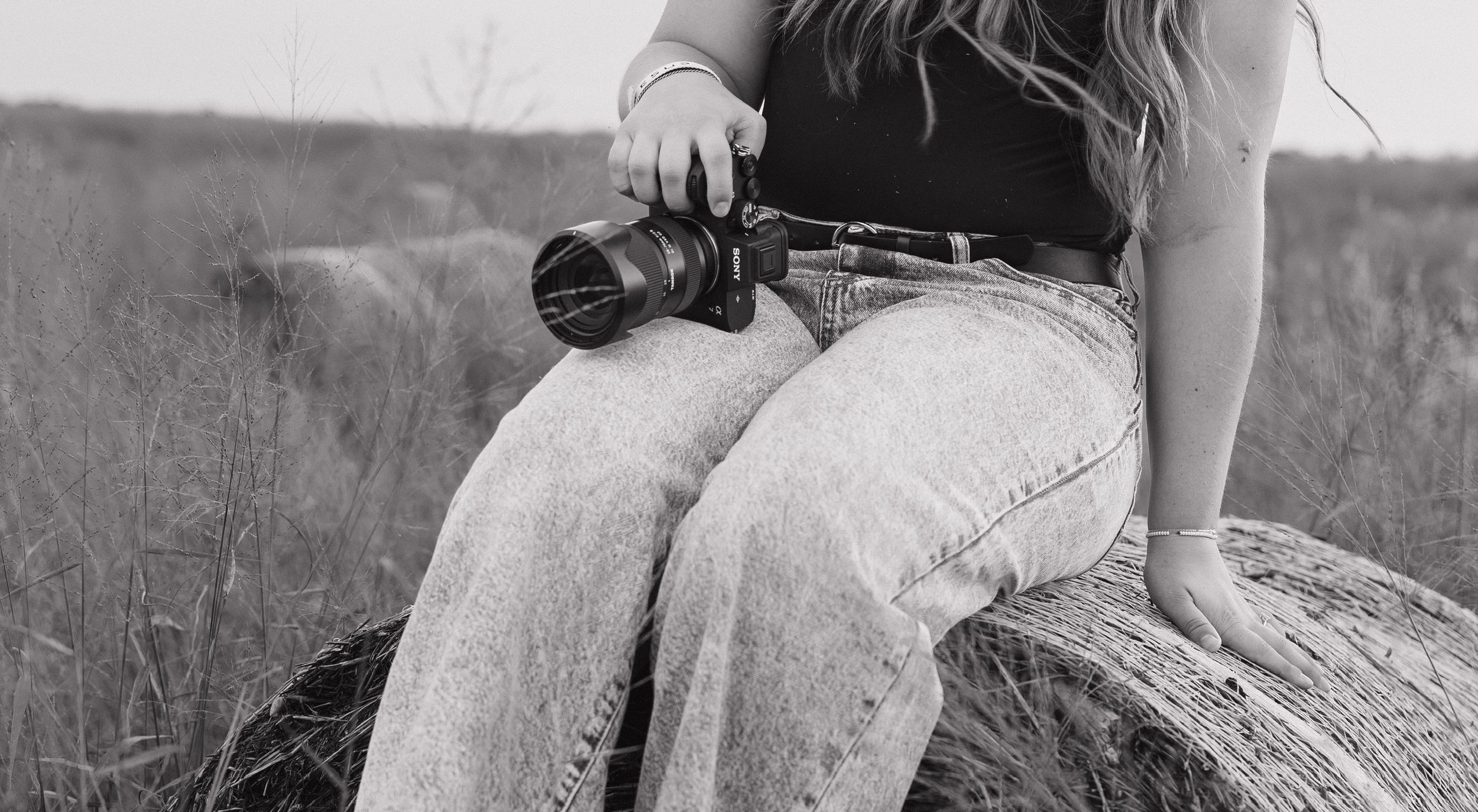 Black and white image of photographer sitting on rock with camera in field.