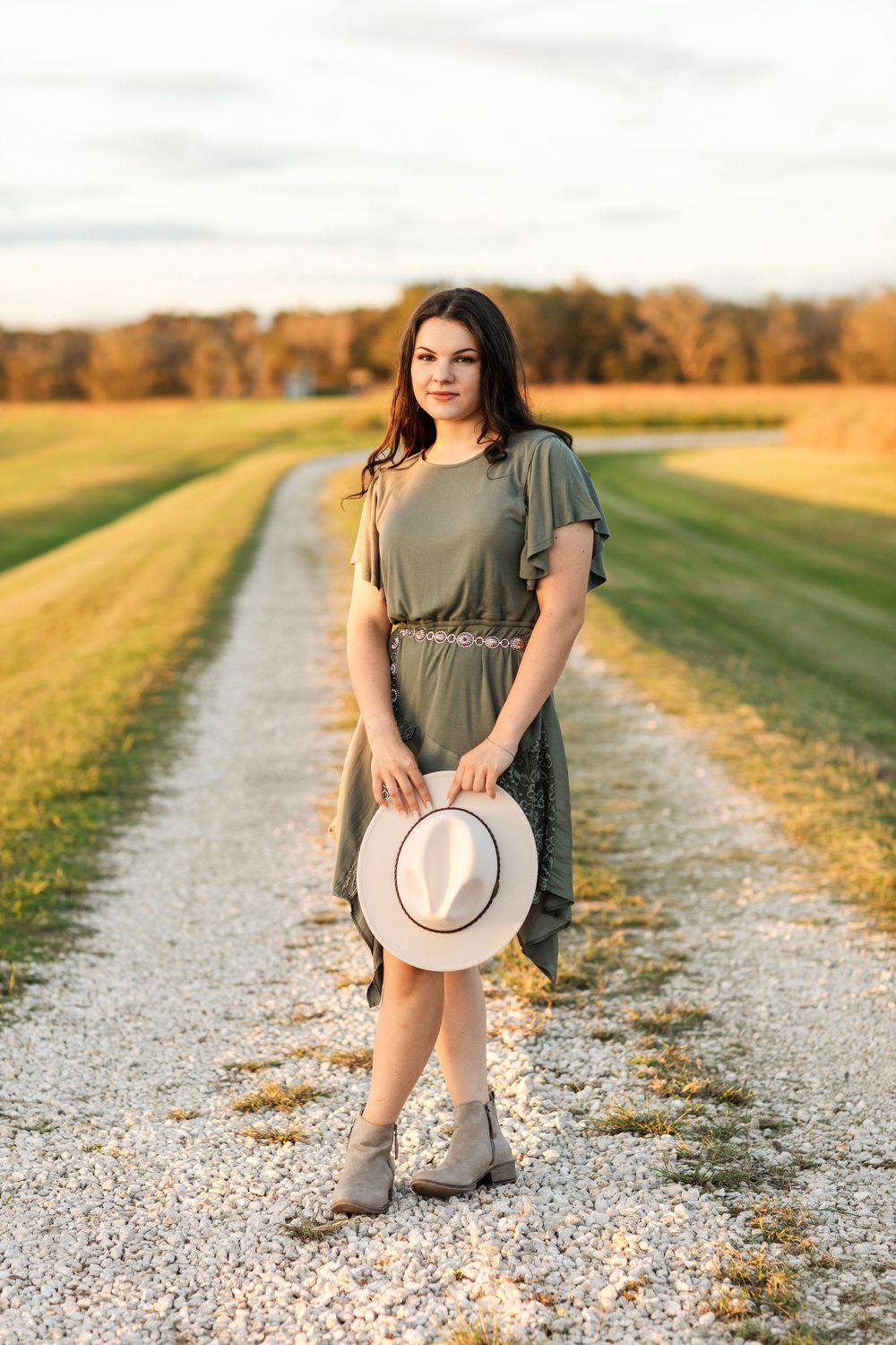A person sits on a gravel path wearing a green dress and boots against a backdrop of autumn fields at sunset.