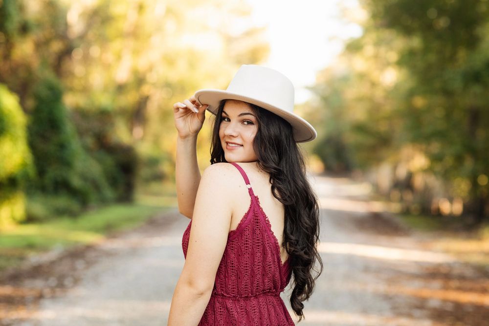 A woman in a burgundy dress and wide-brimmed hat poses on a sunlit pathway surrounded by autumn trees.