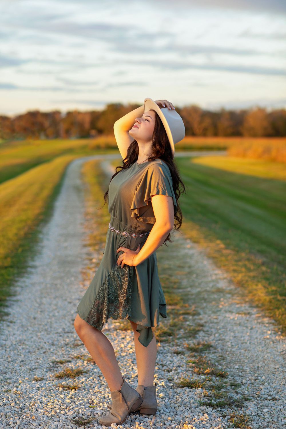 A person in a casual dress and boots strikes a joyful pose on a gravel path during golden hour in autumn.
