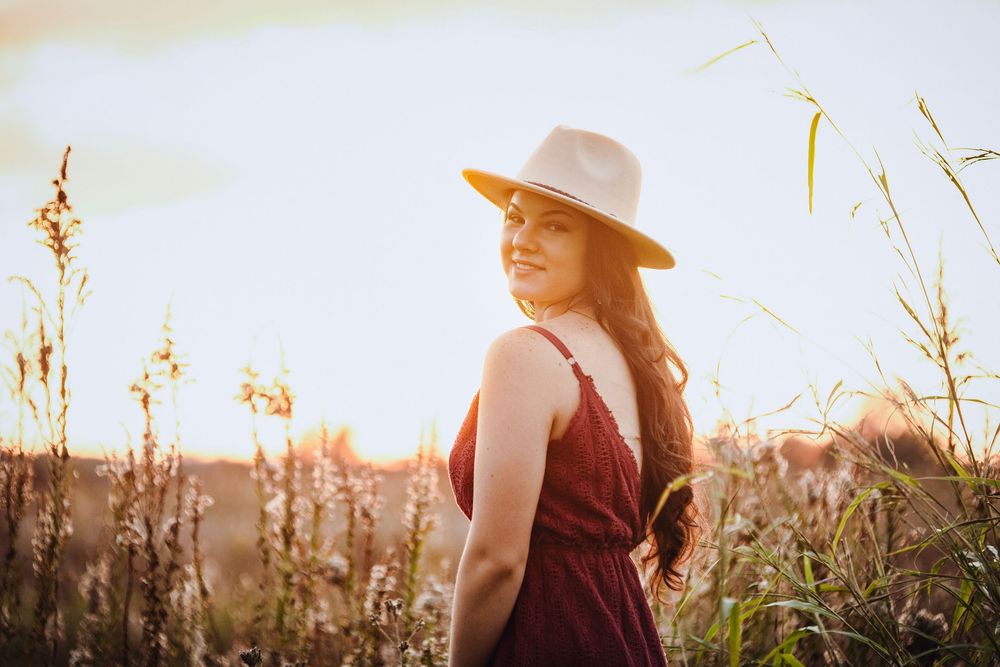 A person in a brown hat and dress walking through tall grass and wildflowers during golden hour sunset.