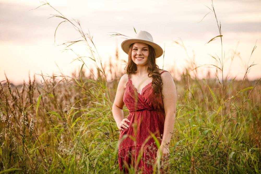 A person in a flowing red dress poses gracefully in a golden field of tall grass at sunset.