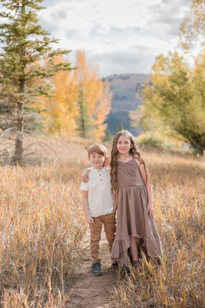 Children in earth-toned clothing stand together in a golden autumn field with mountains and evergreen trees in background.