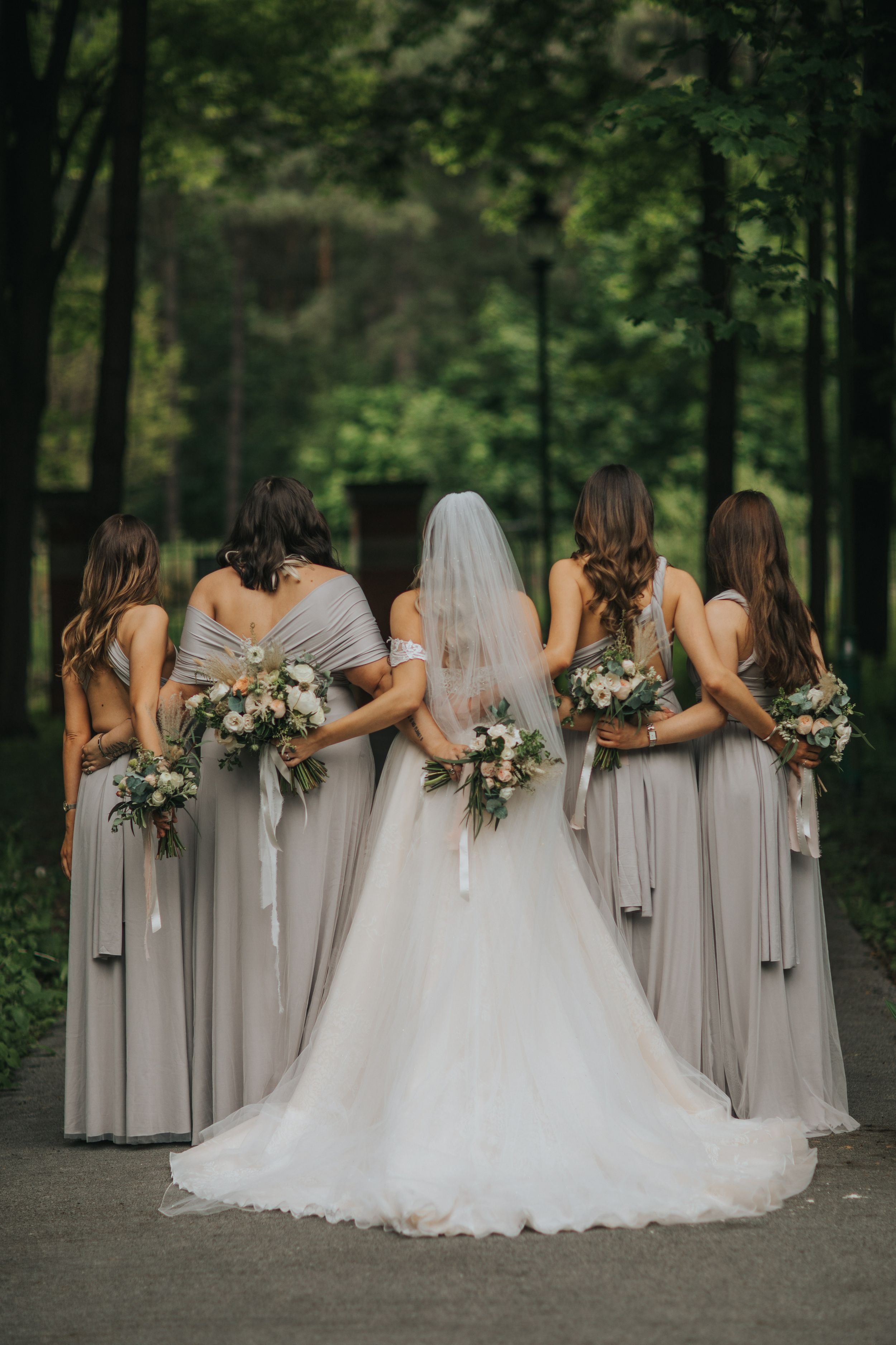 Bride in white gown and bridesmaids in matching gray dresses pose with floral bouquets on a wooded path.