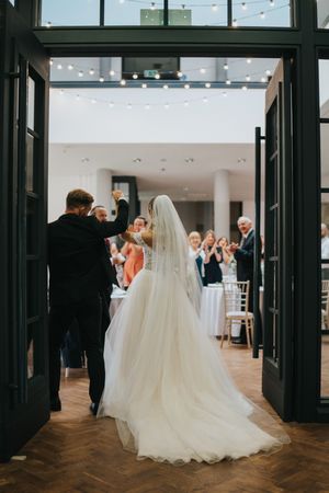 A bride in a flowing white wedding gown enters a venue through large black doors as guests watch from inside.