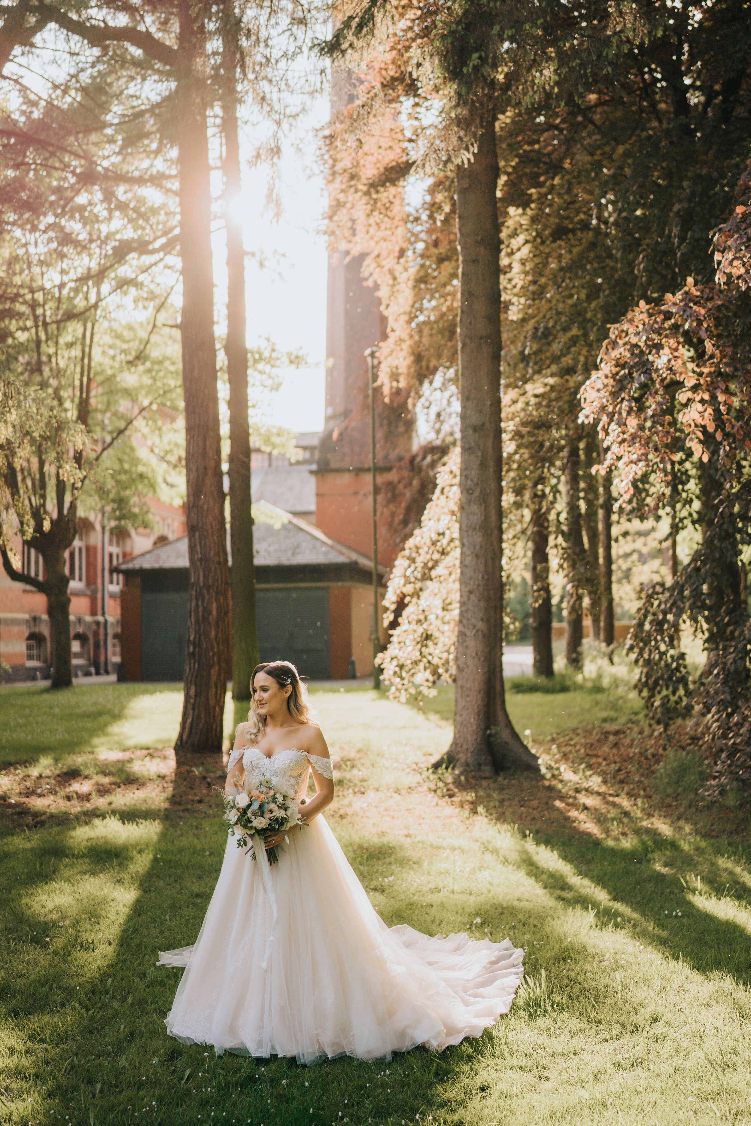 A white wedding dress stands elegantly against tall trees in a sunlit garden with brick buildings in the background.