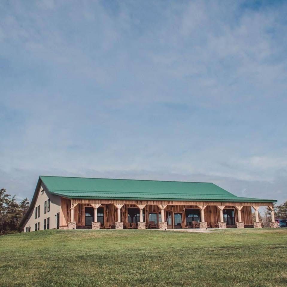 Rustic barn venue with green roof, wooden porch, and stone pillars surrounded by open grassy field under blue sky