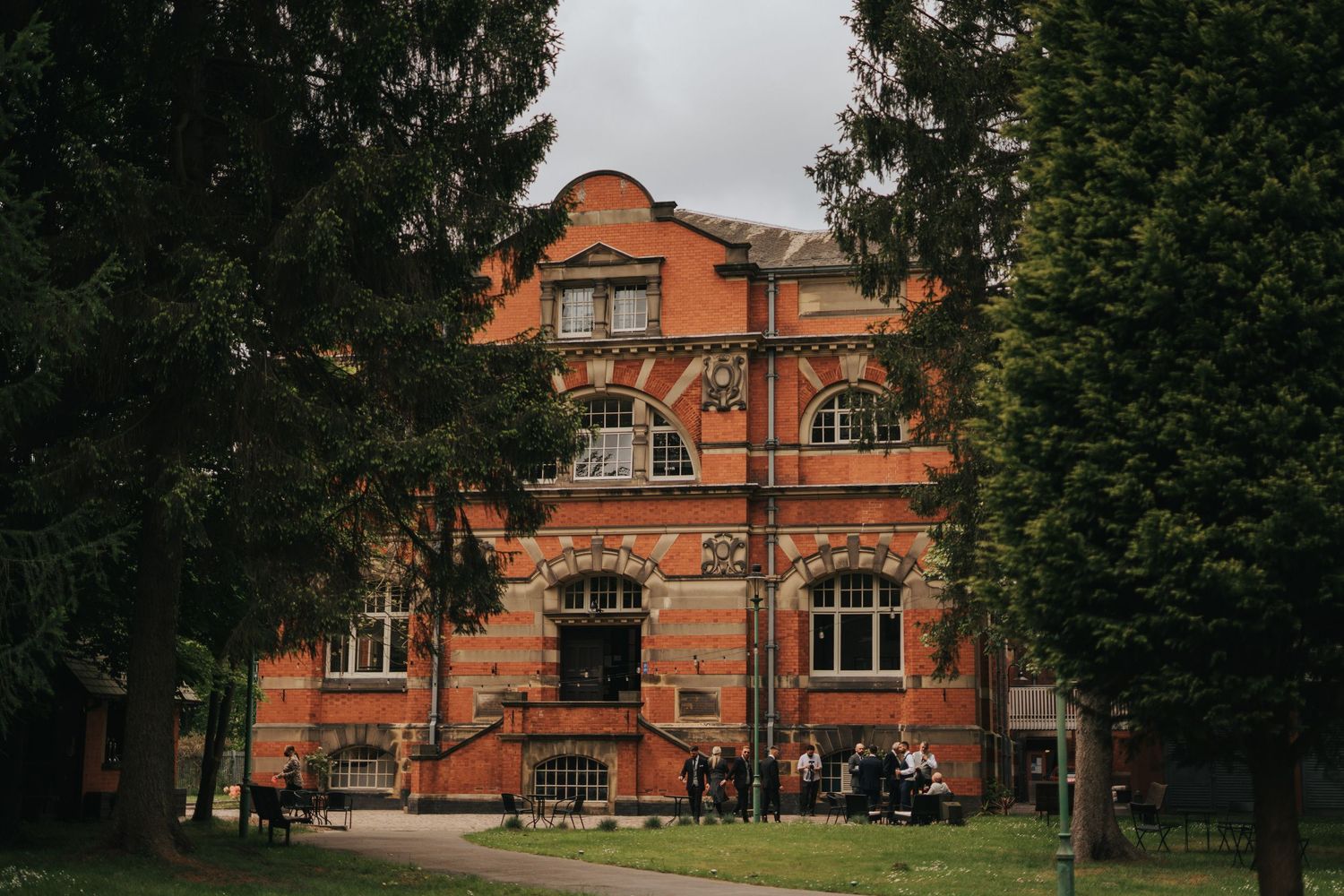 Historic red brick Victorian-style building with ornate architecture framed by evergreen trees on a cloudy day.