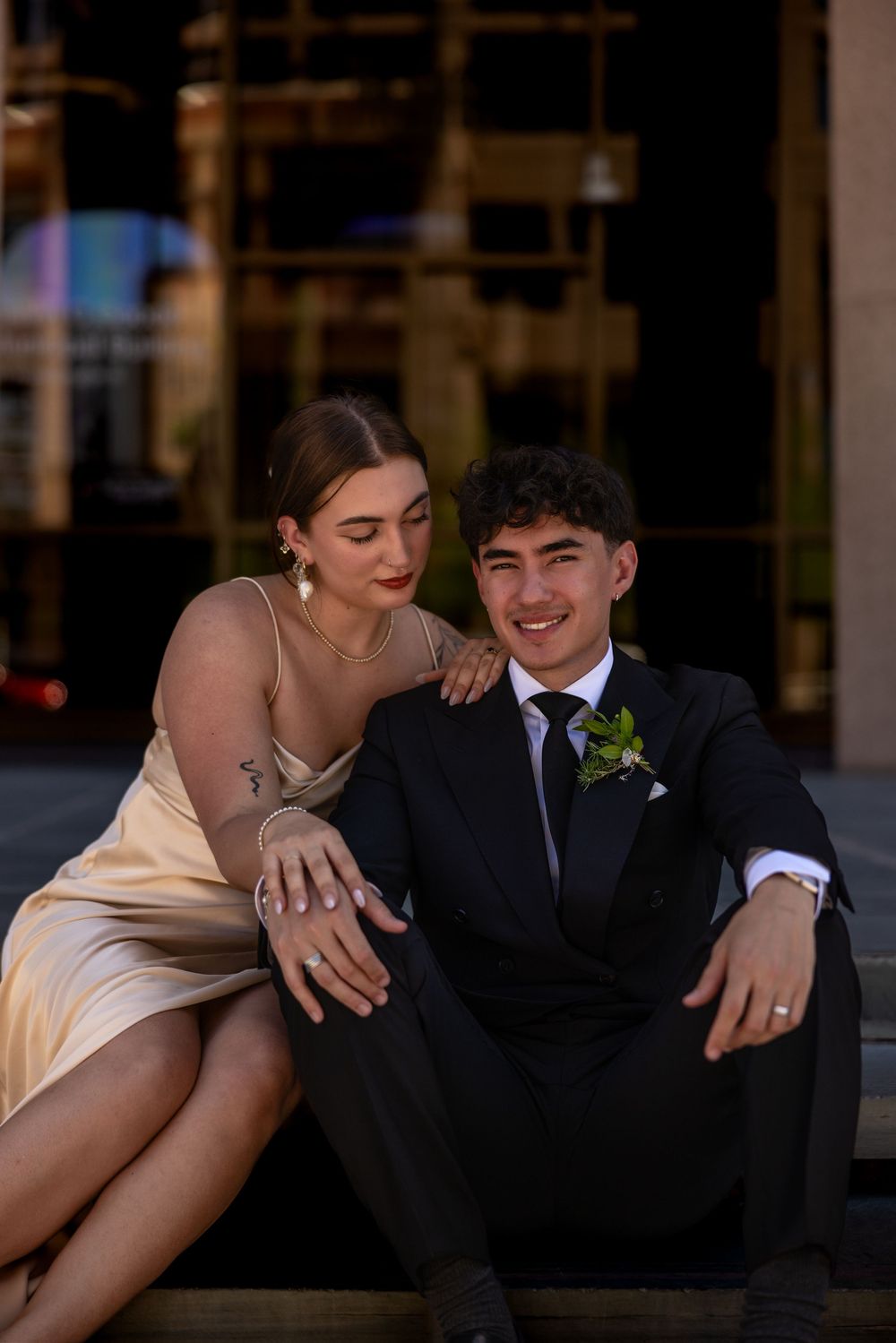 Elegant couple in formal wear sits together outdoors with woman leaning on mans shoulder.