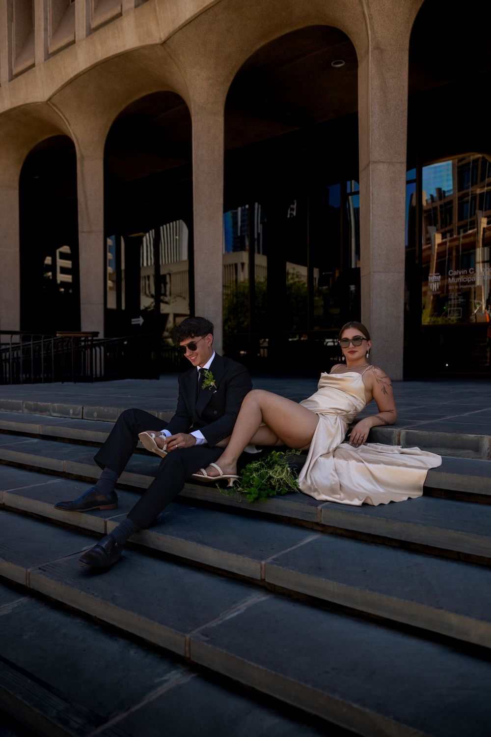 Stylish couple in formal wear poses on urban steps with dramatic arched building backdrop.