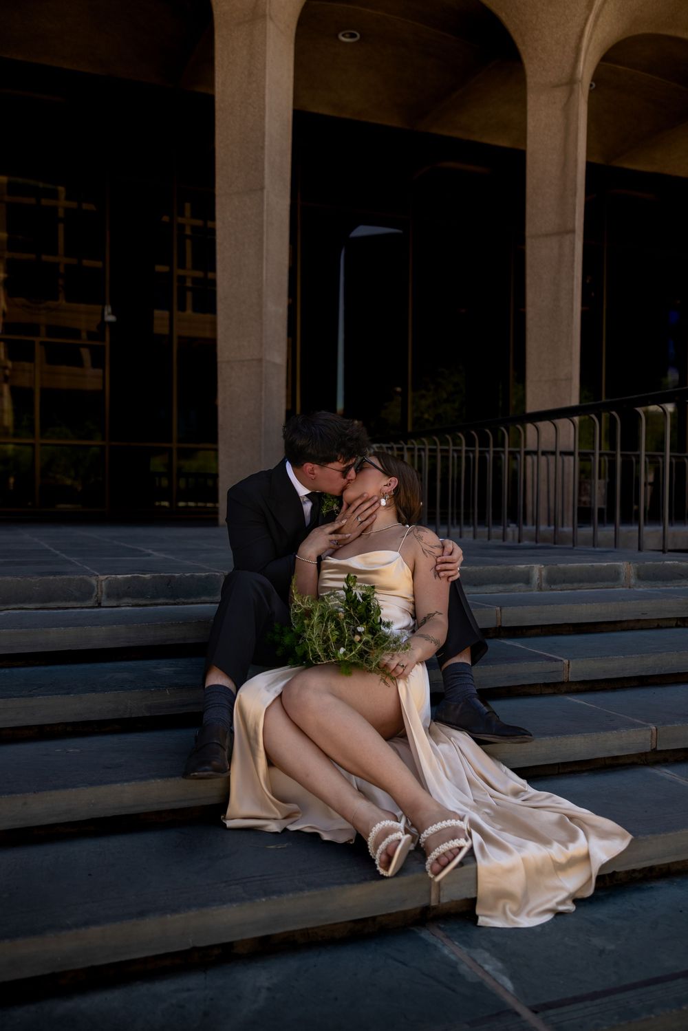 Newlyweds share a passionate kiss while seated on city steps with bouquet of greenery.